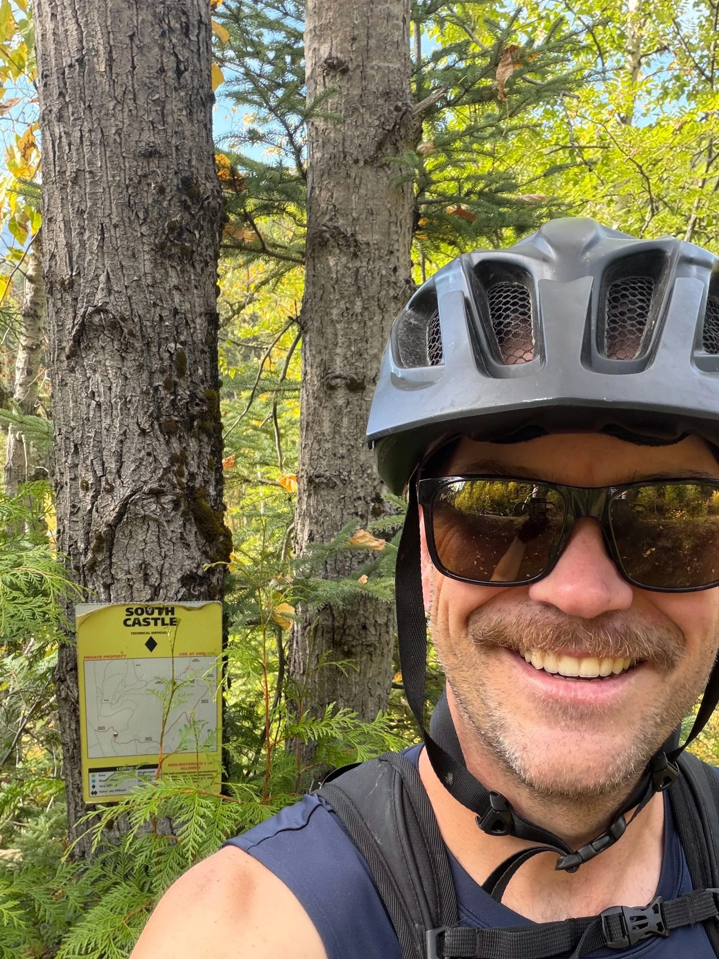 A man taking a selfie outdoors in a forested area, wearing a black helmet and sunglasses, with a bright smile. There is a tree with a sign that reads 'South Castle' in the background.