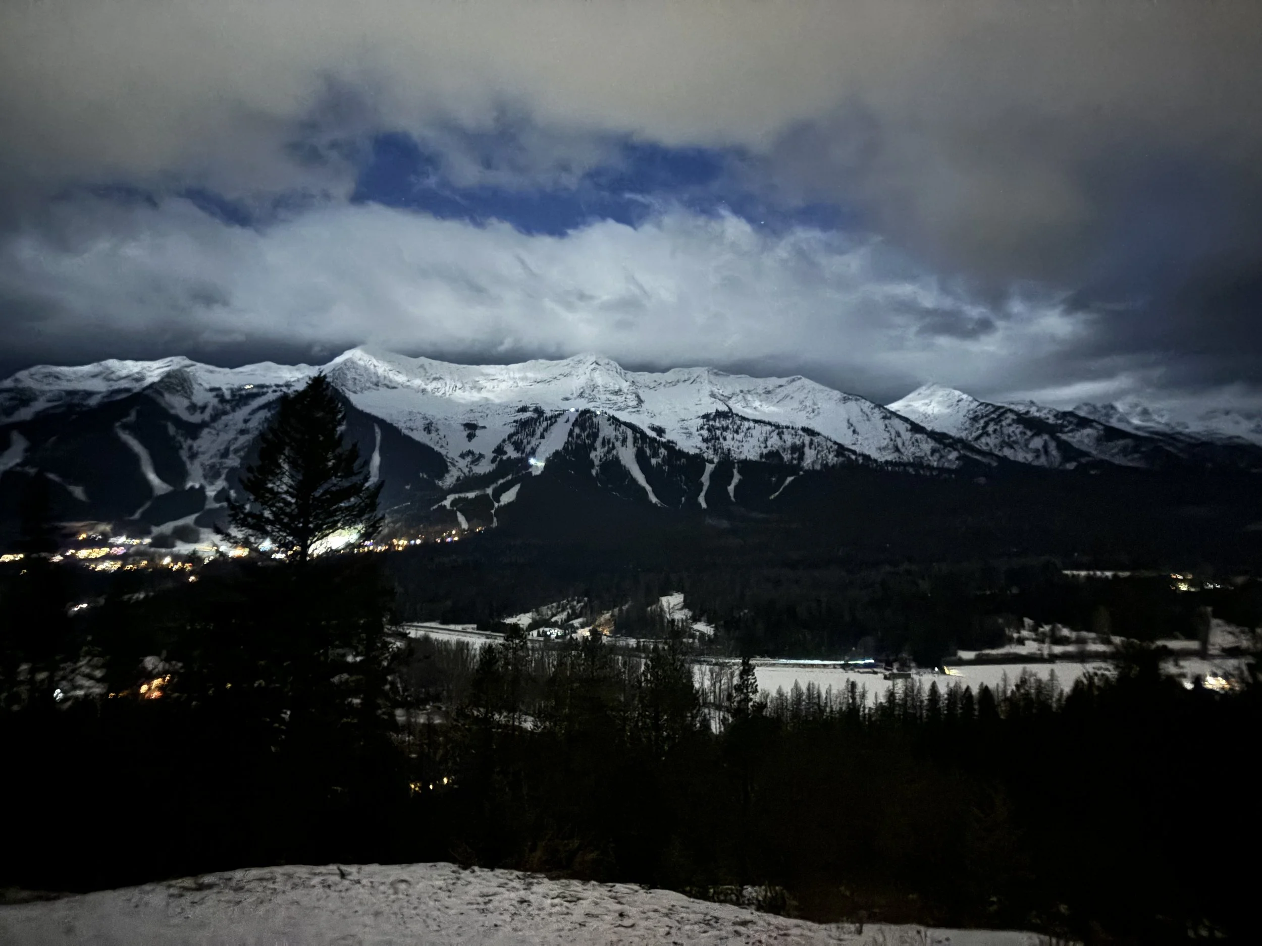 Snow-covered mountain range under dark, cloudy sky at night with a few visible stars and a small village illuminated at the base.