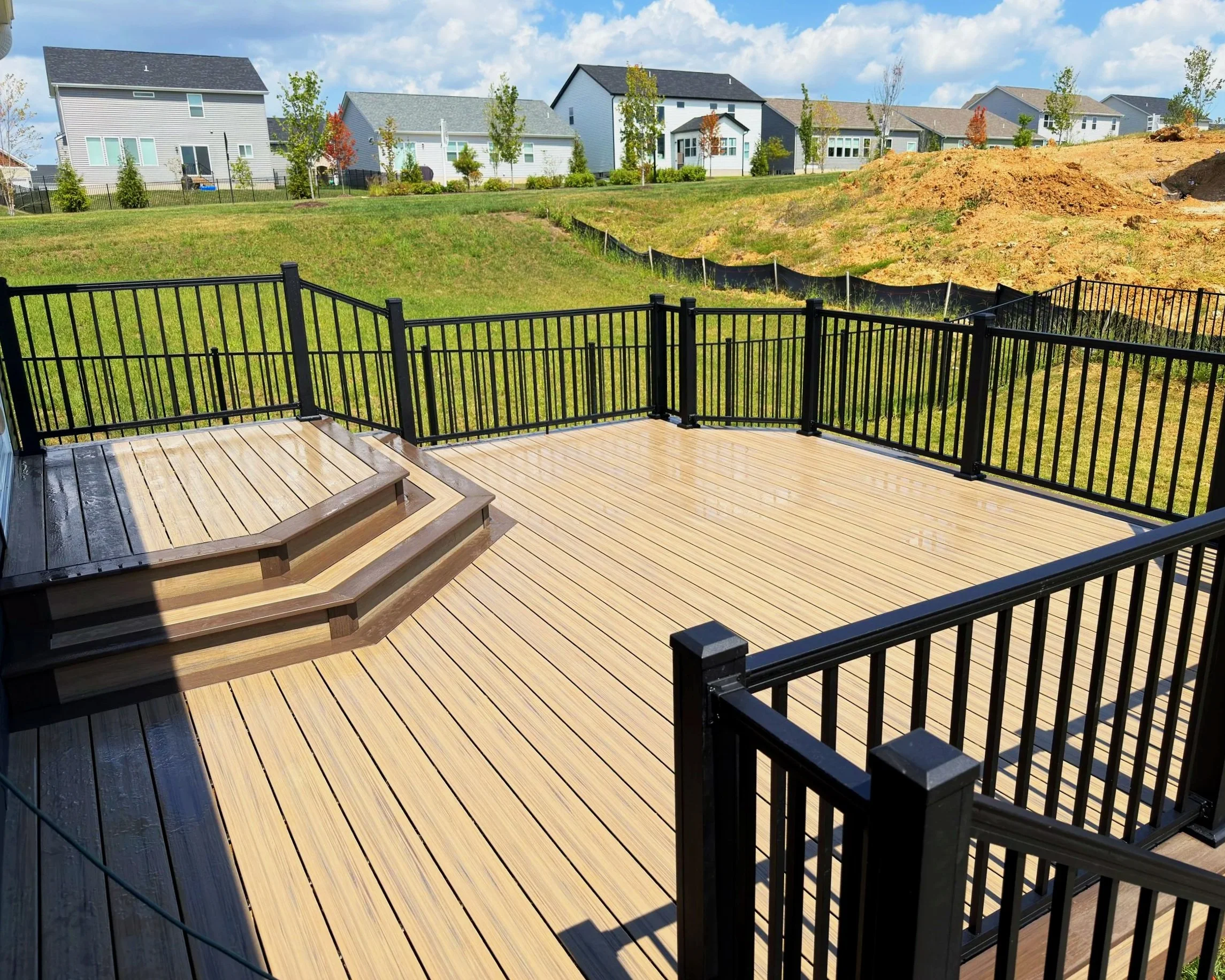 A wooden deck with a black metal railing overlooking a green yard and a row of houses in the background under a partly cloudy sky.