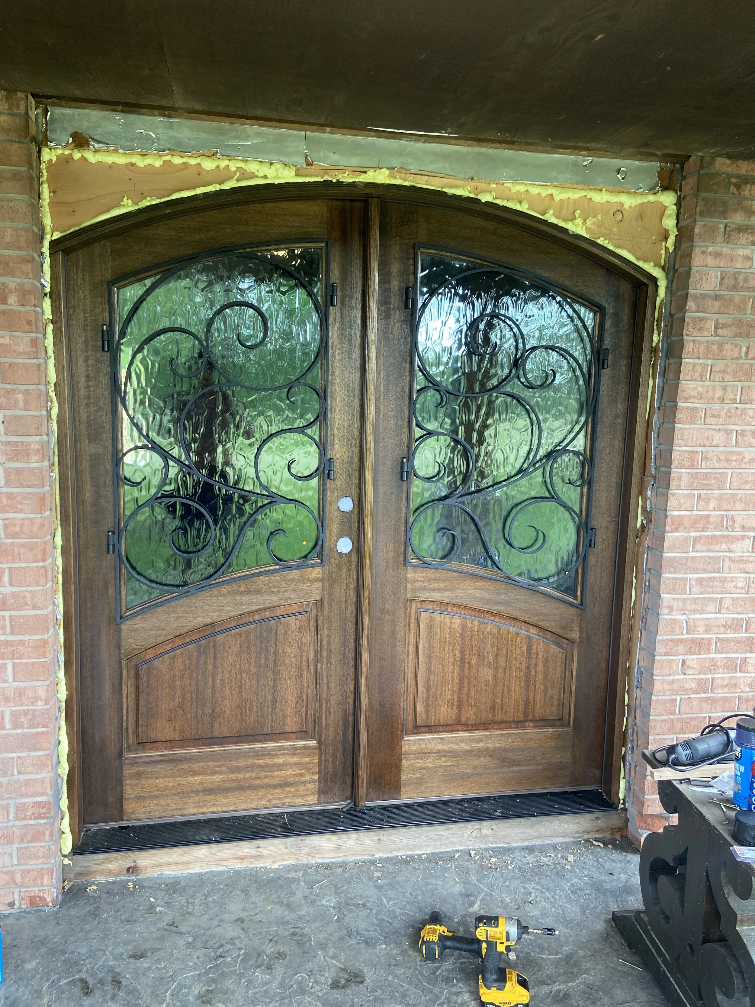 Exterior of a house under renovation showing double wooden front doors with decorative wrought iron grill and glass panels, framed by brick walls, with construction tools and insulation visible.