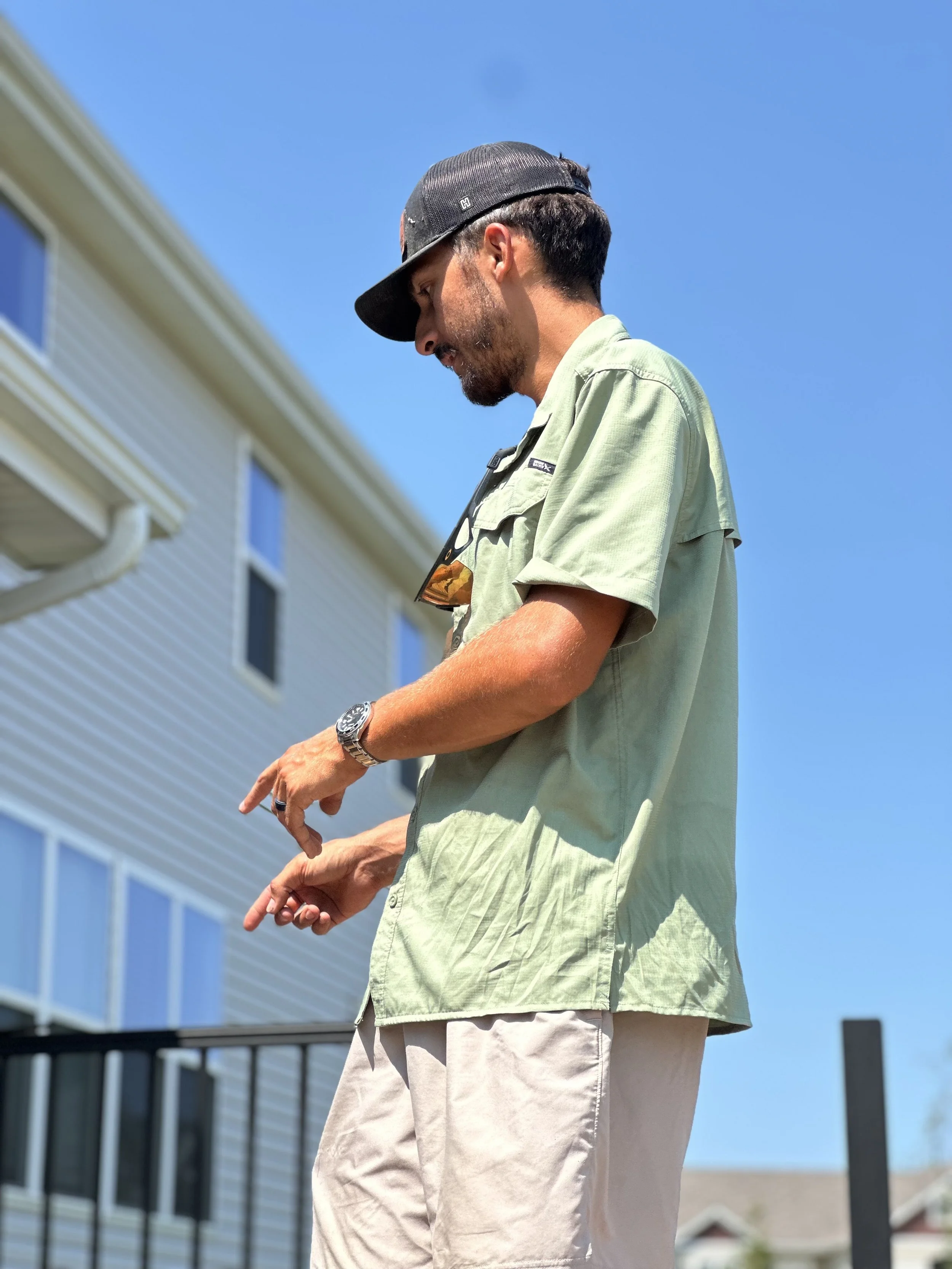 A man with a beard wearing a black baseball cap, light green shirt, and beige pants standing outdoors against a blue sky and a building with windows.