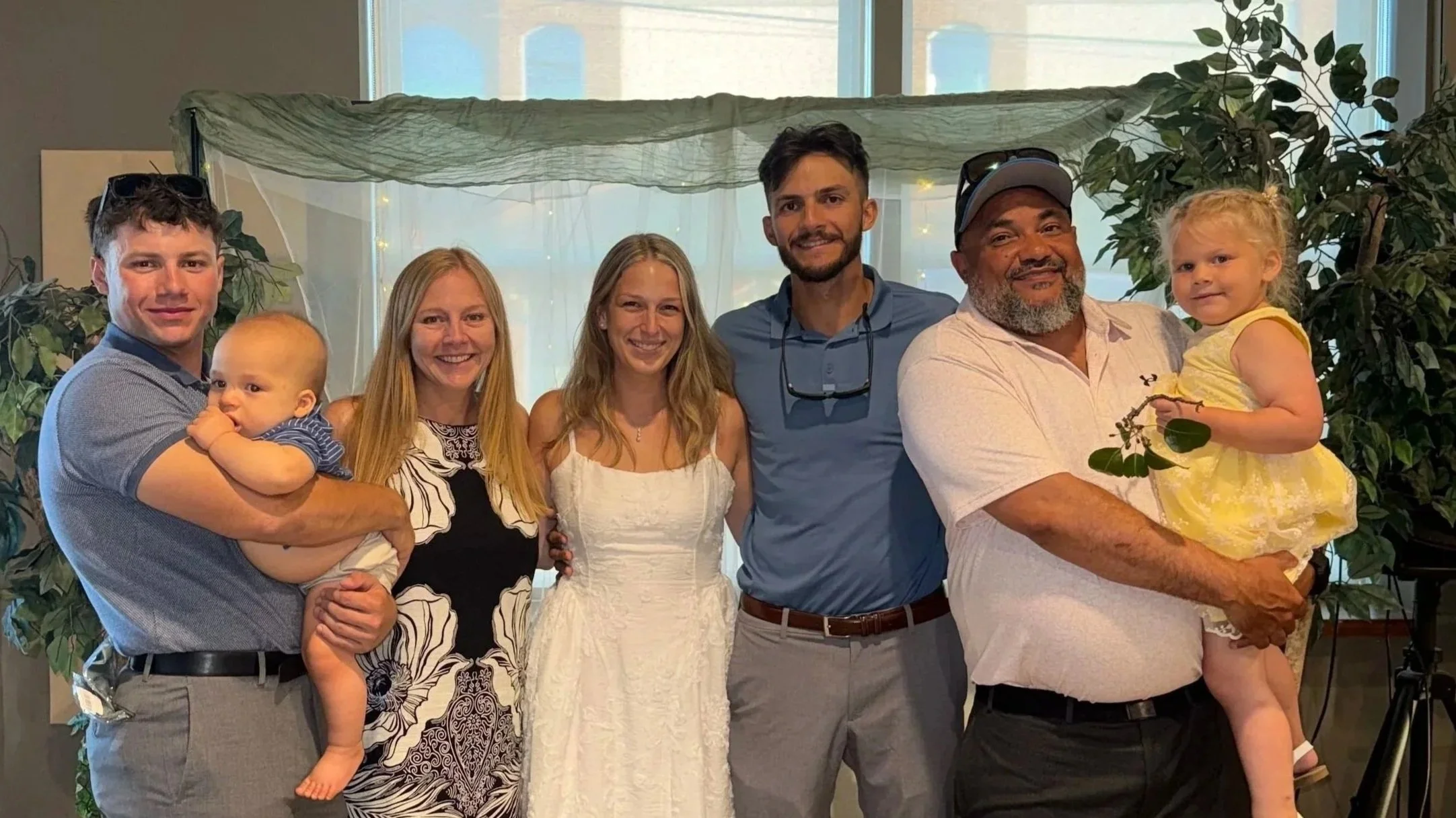 Group of seven people, including adults and children, smiling and posing together indoors, with plants and a window in the background.