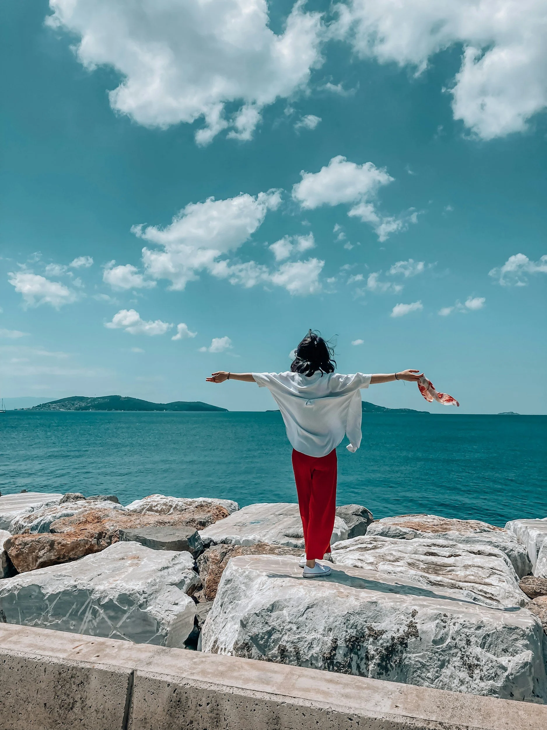 Person standing on rocks by the ocean with arms outstretched, facing the water, under a blue sky with clouds. Nature helps reduce anxiety and cope with anxiety.