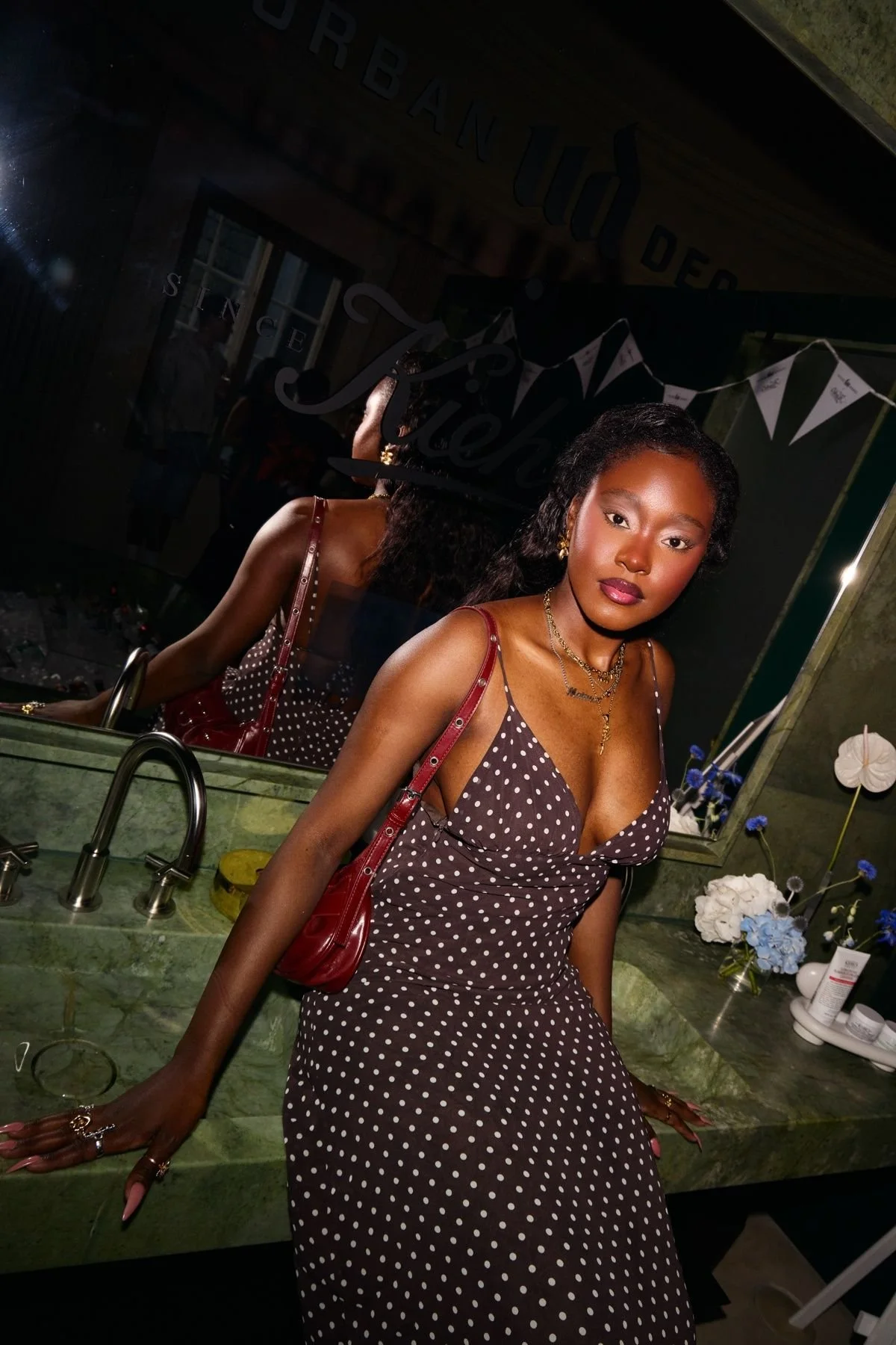 A young woman with curly hair and dark skin taking a selfie in front of a mirror at a sink, wearing a brown dress with white polka dots and gold jewelry, with a red purse on her shoulder. The background includes flowers and a sign that possibly says 'Birthday' and a reflection of her in the mirror.