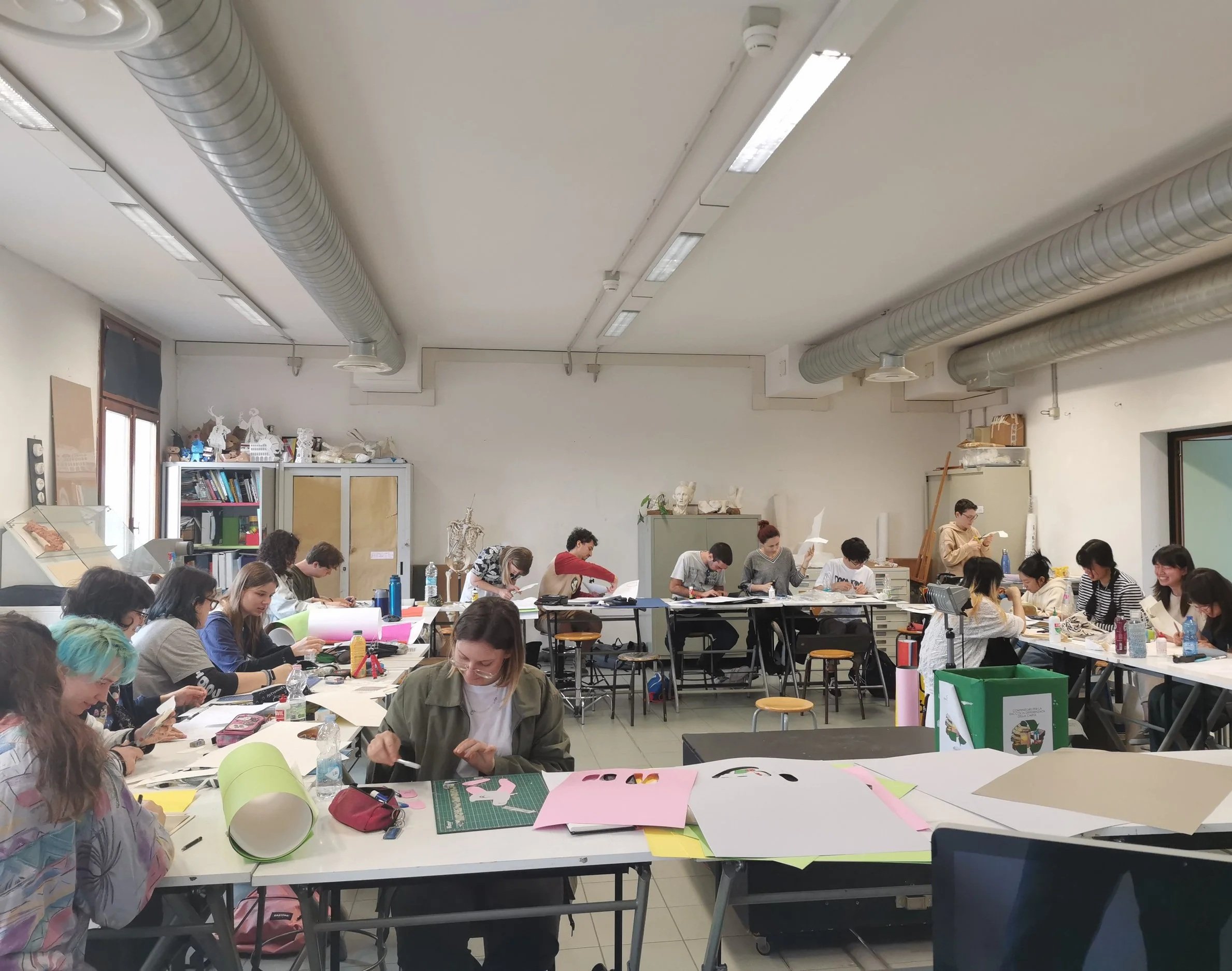 Aula di persone sedute a tavoli che lavorano su progetti artigianali o hobby, in un ambiente spazioso con soffitto alto, tubi di ventilazione visibili e scaffali con libri ed eventualmente materiali decorativi.