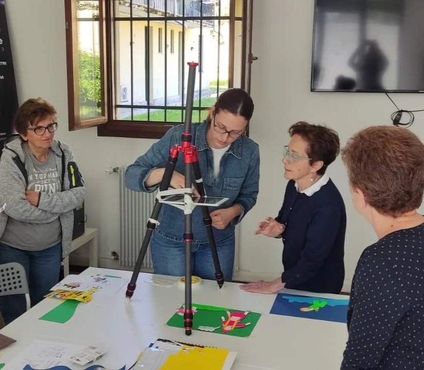 Quattro donne in una stanza discutono intorno a una tavola con vari pezzi di cartone colorati e una macchina fotografica su treppiede.
