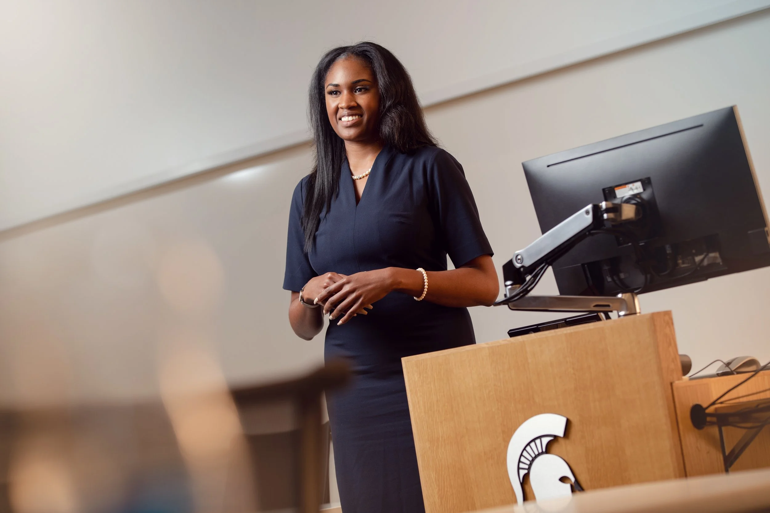 A woman with long black hair wearing a dark navy dress and pearl necklace standing at a lectern with the Michigan State University logo, speaking or presenting in a classroom or conference room.