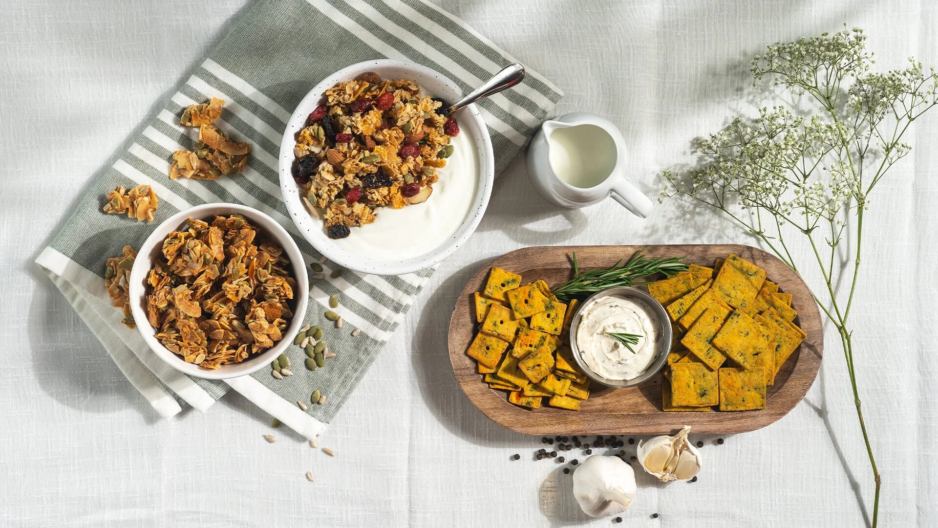 A flat lay of Indian vegetarian snacks and ingredients on a white tablecloth. Includes bowls of granola, crispy snack bites, a wooden platter of turmeric and spinach squares with a dipping sauce, garlic, and black peppercorns, with green herbs and a green-and-white striped cloth.