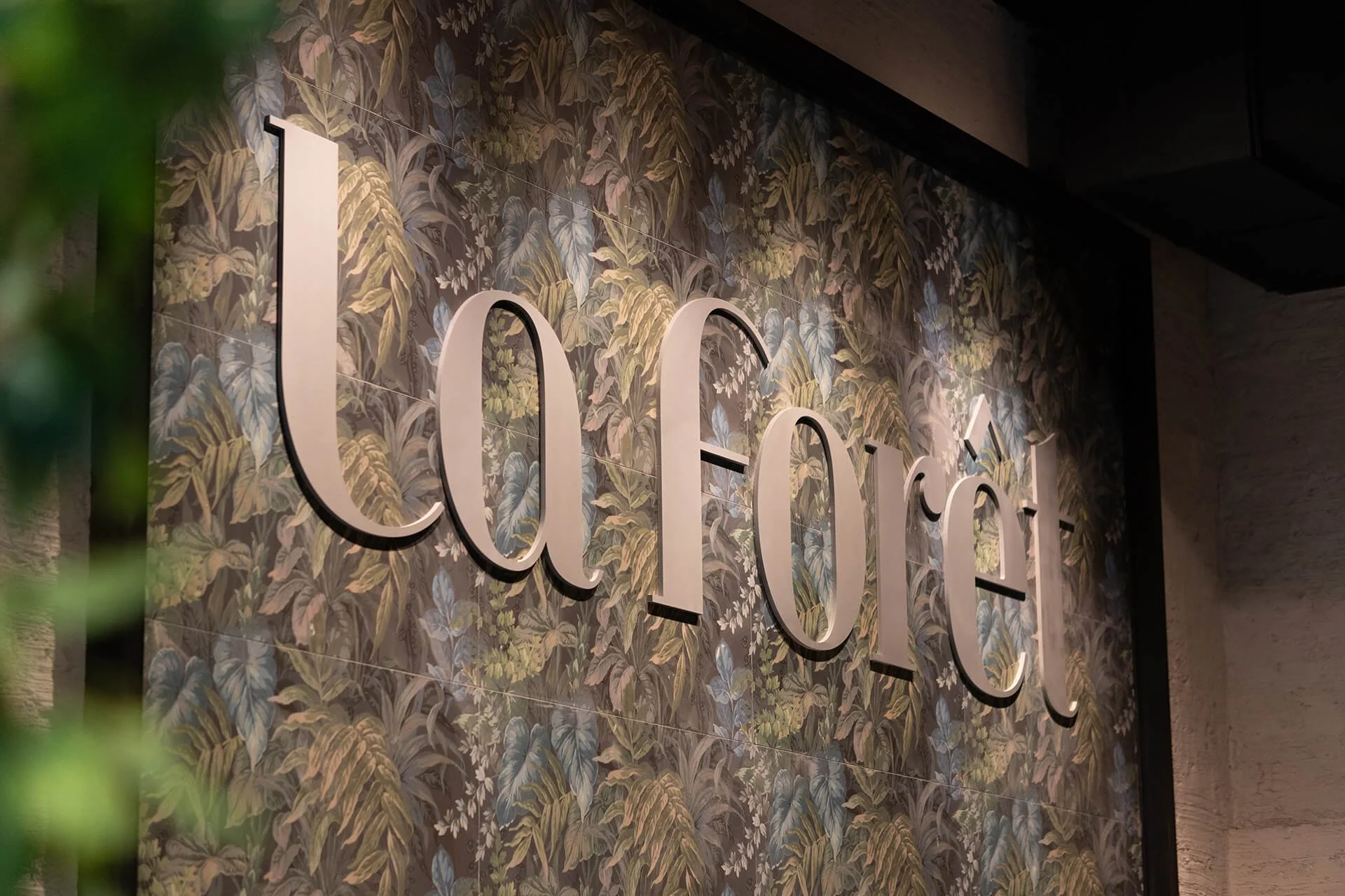 Close-up of a wall with a leafy, tropical-themed wallpaper and white 3D letters spelling 'COCO' mounted on it.