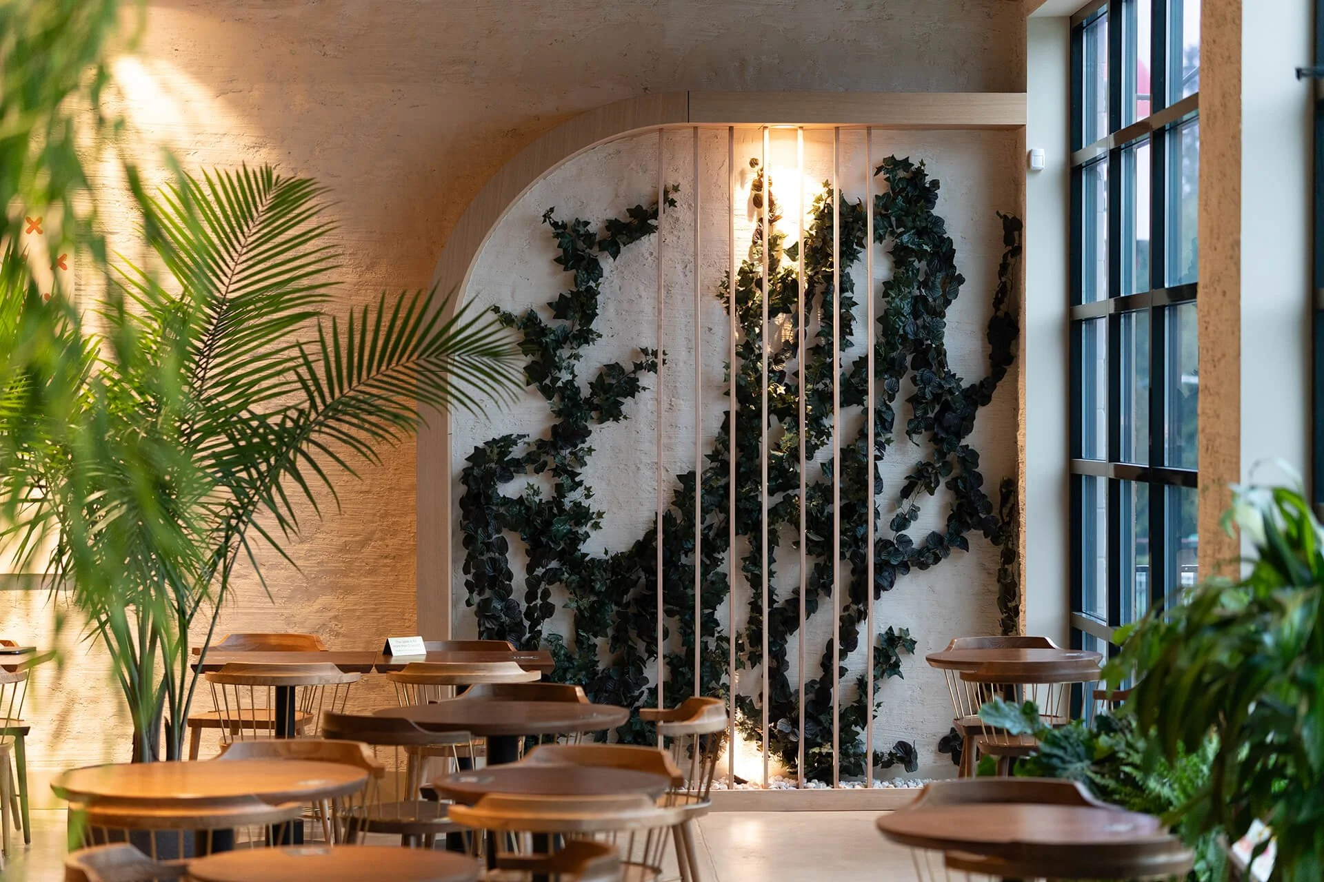 Interior of a modern cafe or restaurant with wooden tables and chairs, decorated with green plants and a wall featuring climbing ivy and vertical wooden slats. Large windows let in natural light.