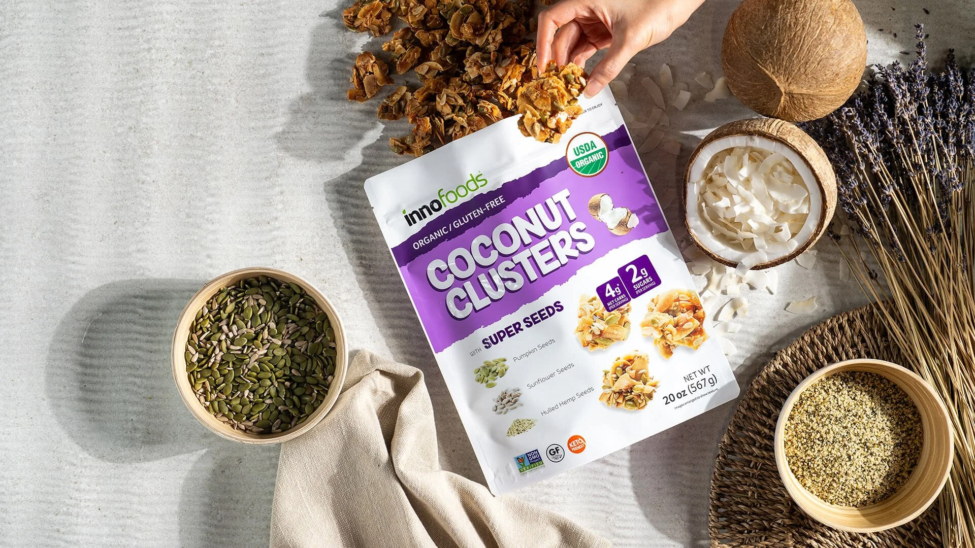 A bag of Inno Foods organic, gluten-free coconut clusters with super seeds, including pumpkin, sunflower, and hemp seeds, surrounded by bowls of seeds, coconut, and dried herbs on a light gray surface.