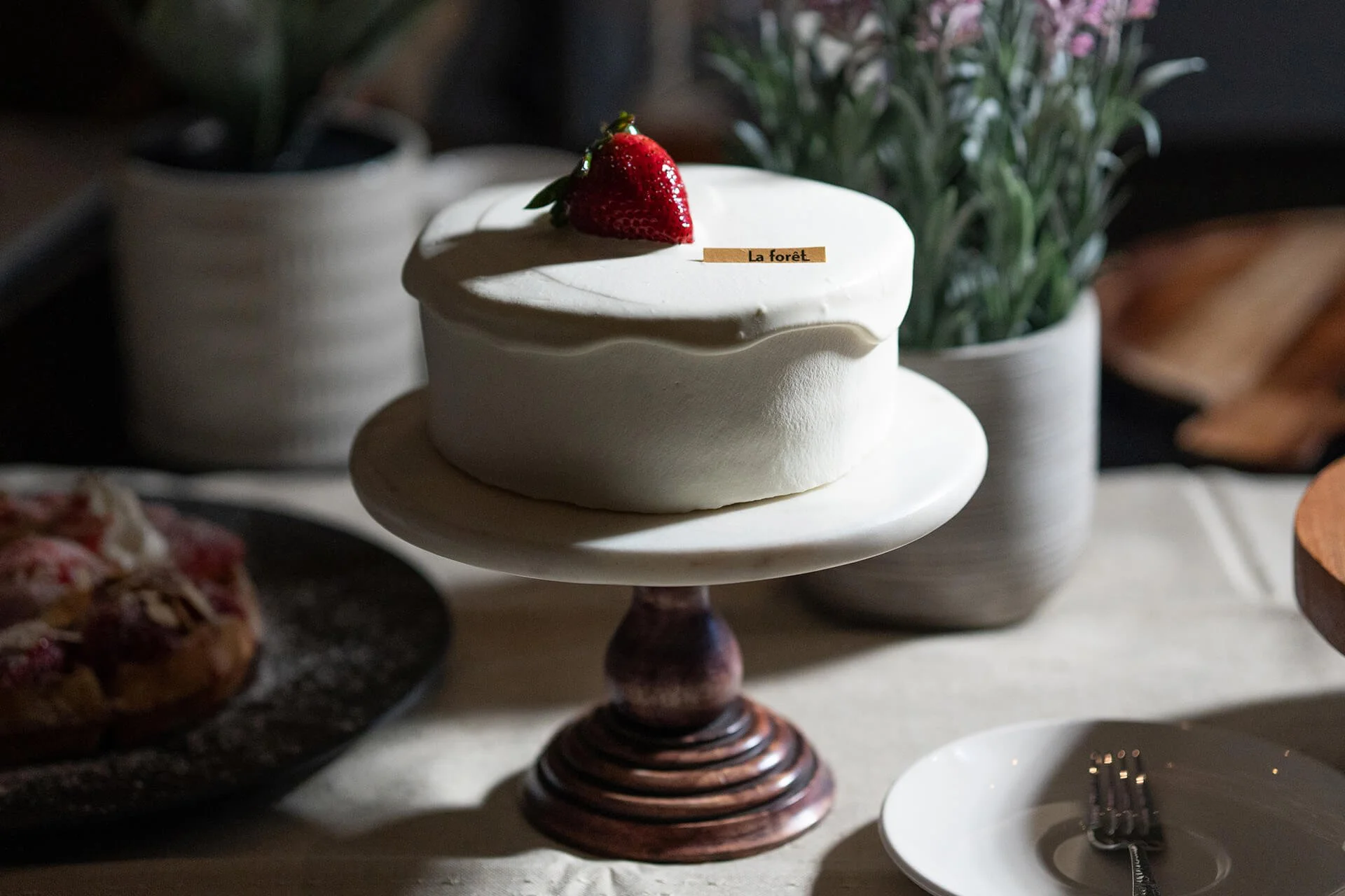 A small round cake with white frosting topped with a fresh strawberry, placed on a white cake stand. The cake has a label that reads 'La forêt.' In the background, there are green plants and other desserts visible.
