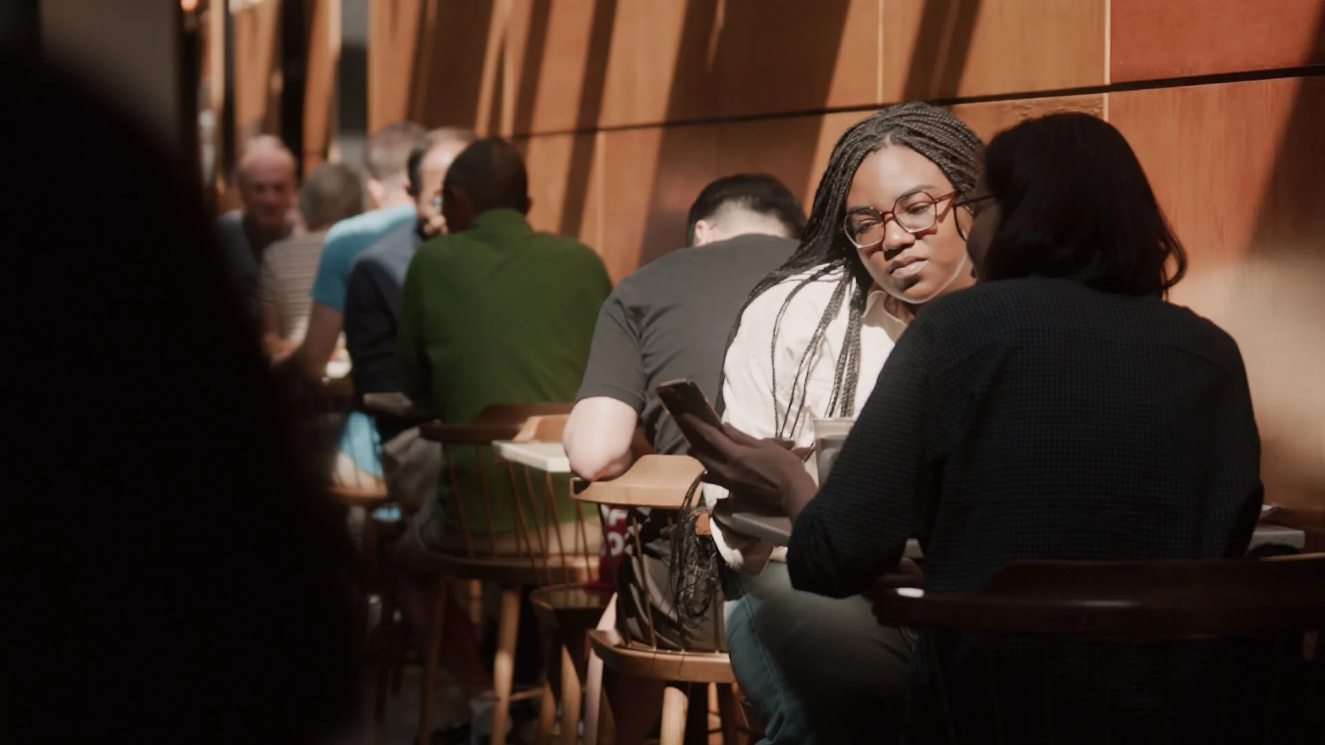 A group of diverse people sitting closely together at a long table, engaging in conversation and looking at their phones in a cozy indoor setting with wooden paneling.