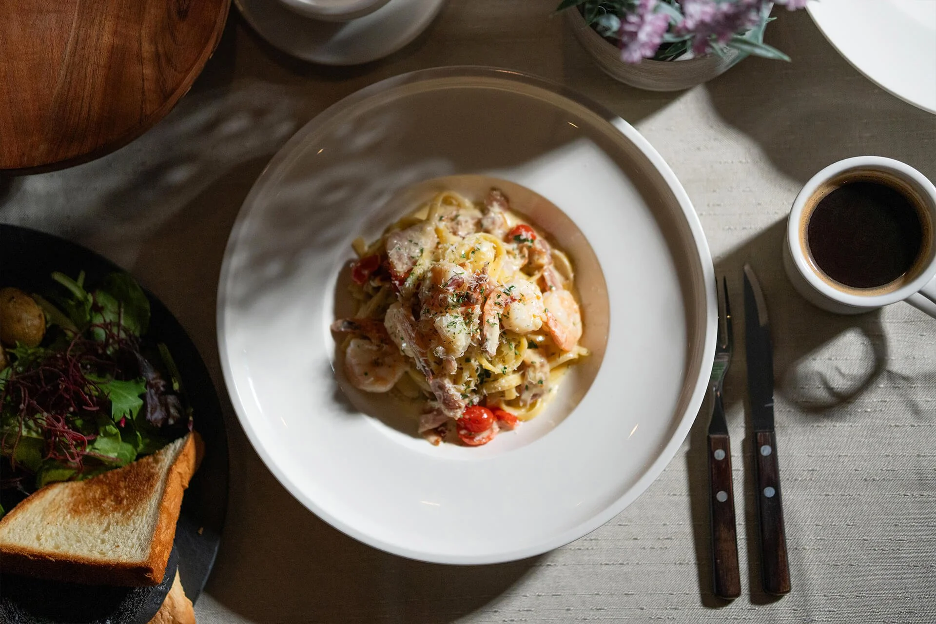 A plate of creamy pasta with shrimp and cherry tomatoes, served with a cup of coffee and a side salad on a black plate with toast, on a table with utensils and a flower vase.
