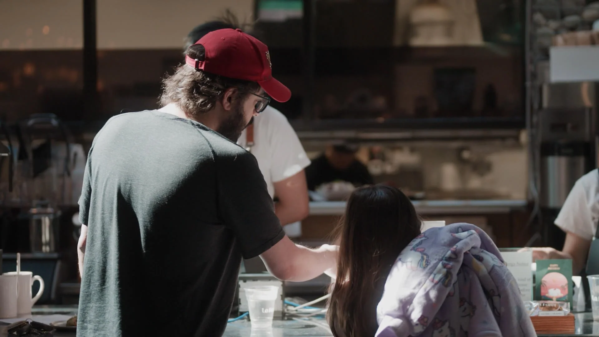 A man with glasses, a beard, and a red cap is leaning over a table with a young girl in a hoodie, working together on a project in a busy background, likely a classroom or workshop.