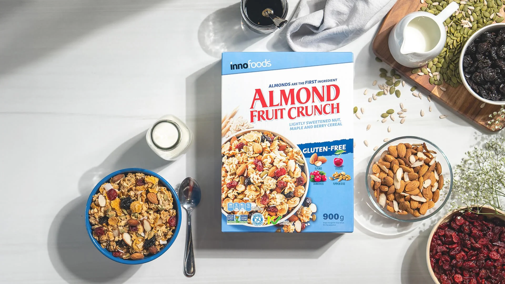 A box of almond fruit crunch cereal surrounded by bowls of cereal with dried fruits and nuts, a spoon, a glass of black coffee, a small pitcher of milk, and scattered pumpkin seeds on a white table.