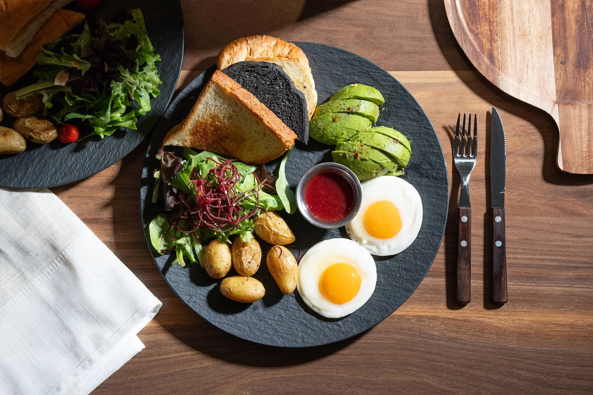 A breakfast plate with eggs, sliced avocado, roasted potatoes, toast, salad greens, and a small container of red sauce on a black plate on a wooden table.