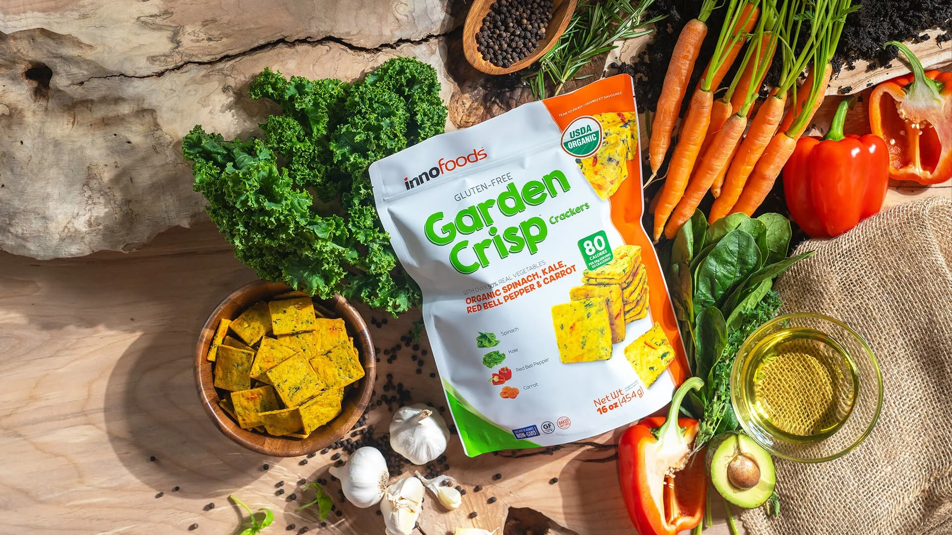A variety of vegetables including fresh carrots, red bell pepper, garlic, and leafy greens, displayed around a package of gluten-free vegetable crackers, with some crackers on a small bowl and a glass of oil or dressing nearby, on a wooden surface with a large rock in the background.