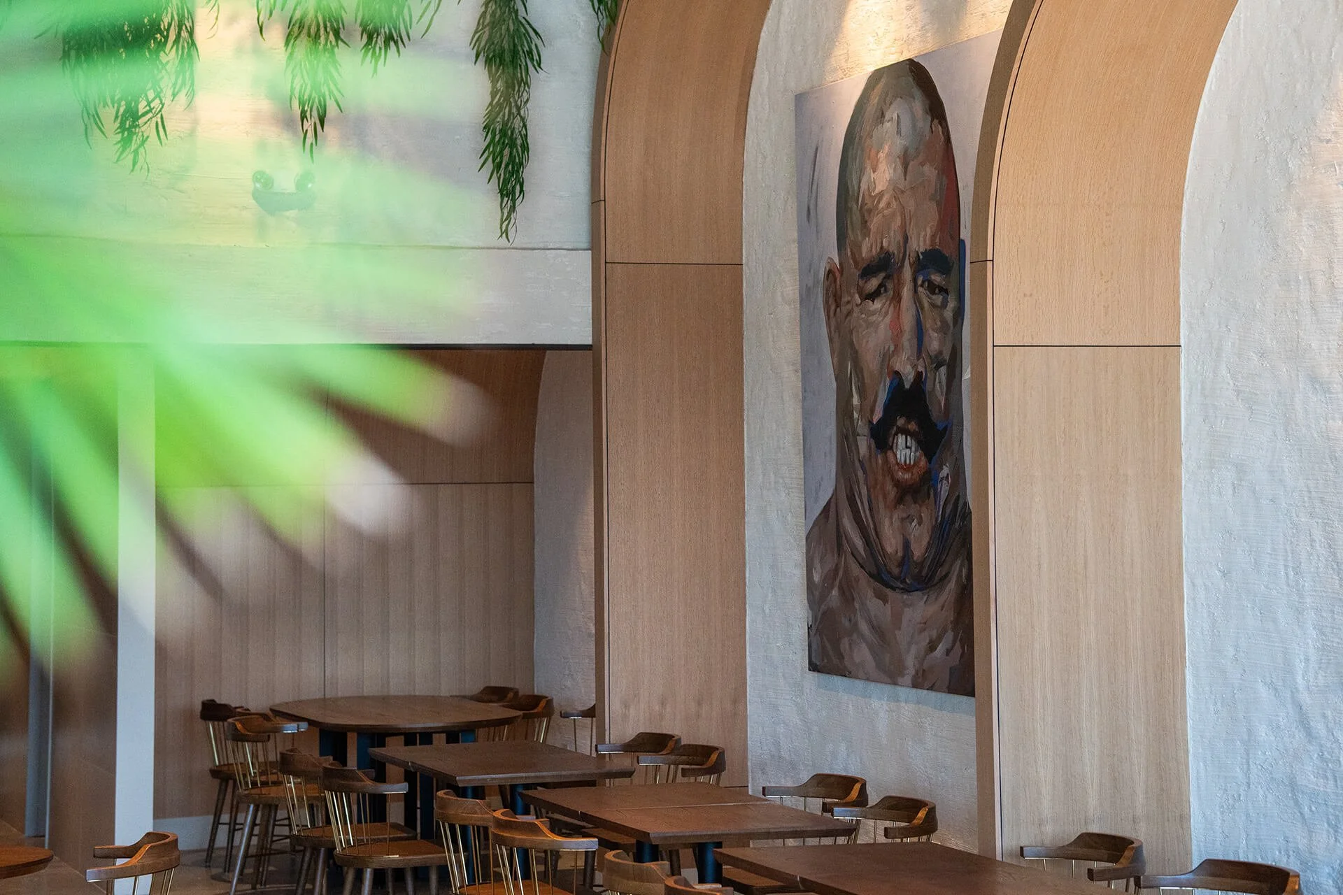 Interior of a modern restaurant or cafe with wooden tables and chairs, a large abstract portrait of a man's face on the wall, and greenery hanging from the ceiling.