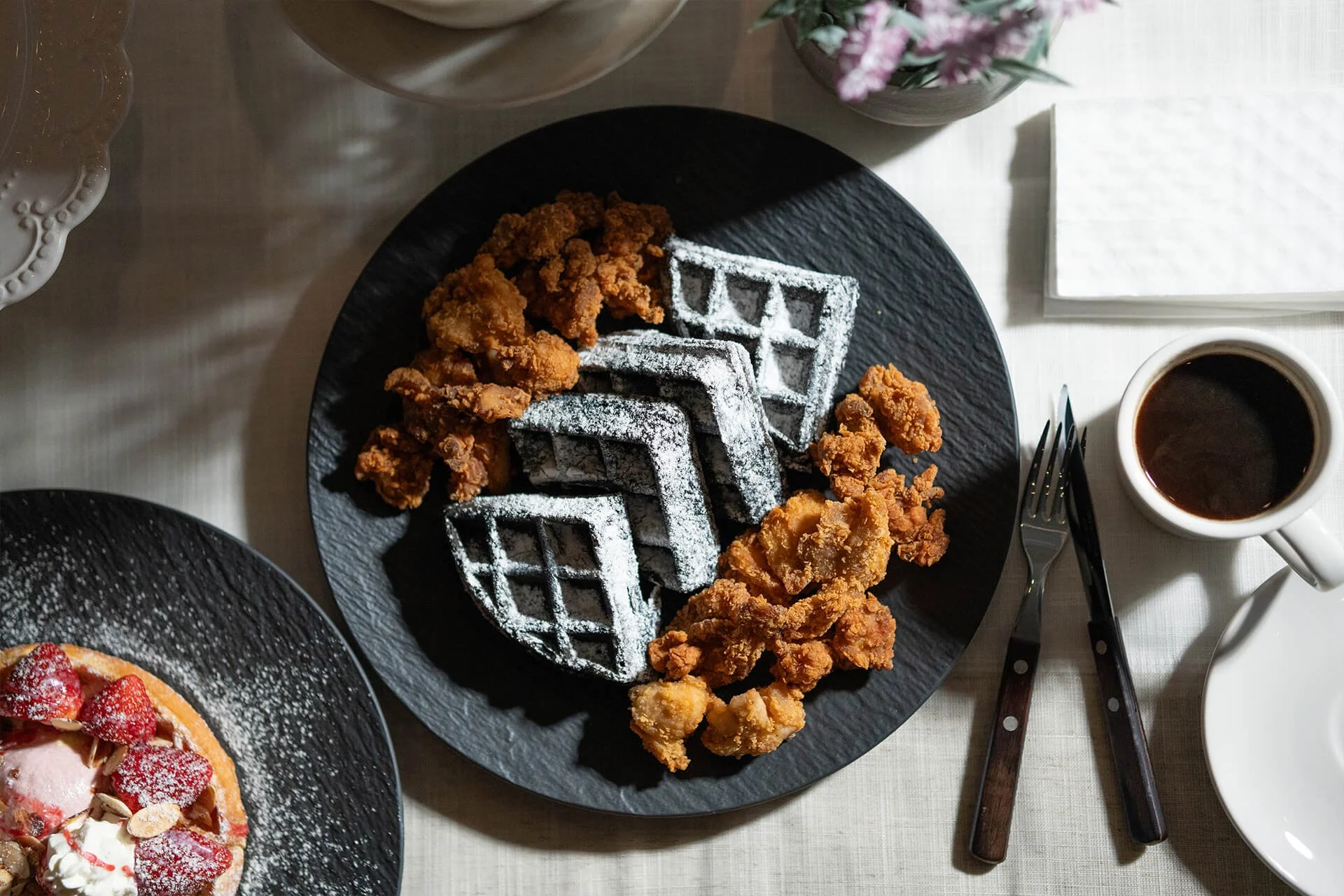 Fried chicken, Belgian waffles dusted with powdered sugar, and a cup of coffee on a white table.