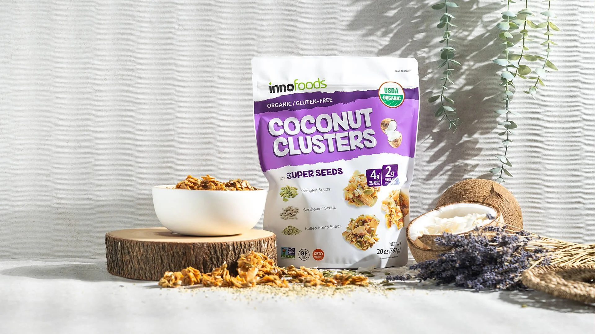 A bag of innofoods organic coconut clusters super seeds, with a bowl of granola in front, placed on a wooden slice, alongside half a coconut and lavender flowers, on a textured background.