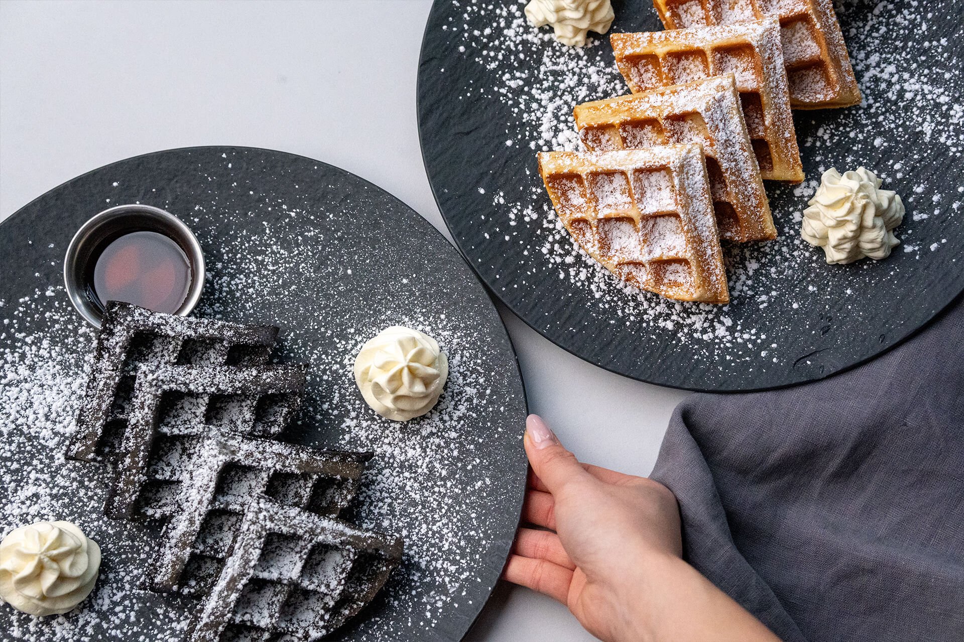 Two black plates with waffles topped with powdered sugar, whipped cream, and syrup. One hand of a person is steadying a plate, and two dollops of whipped cream are on each plate, with powdered sugar dusted over everything.