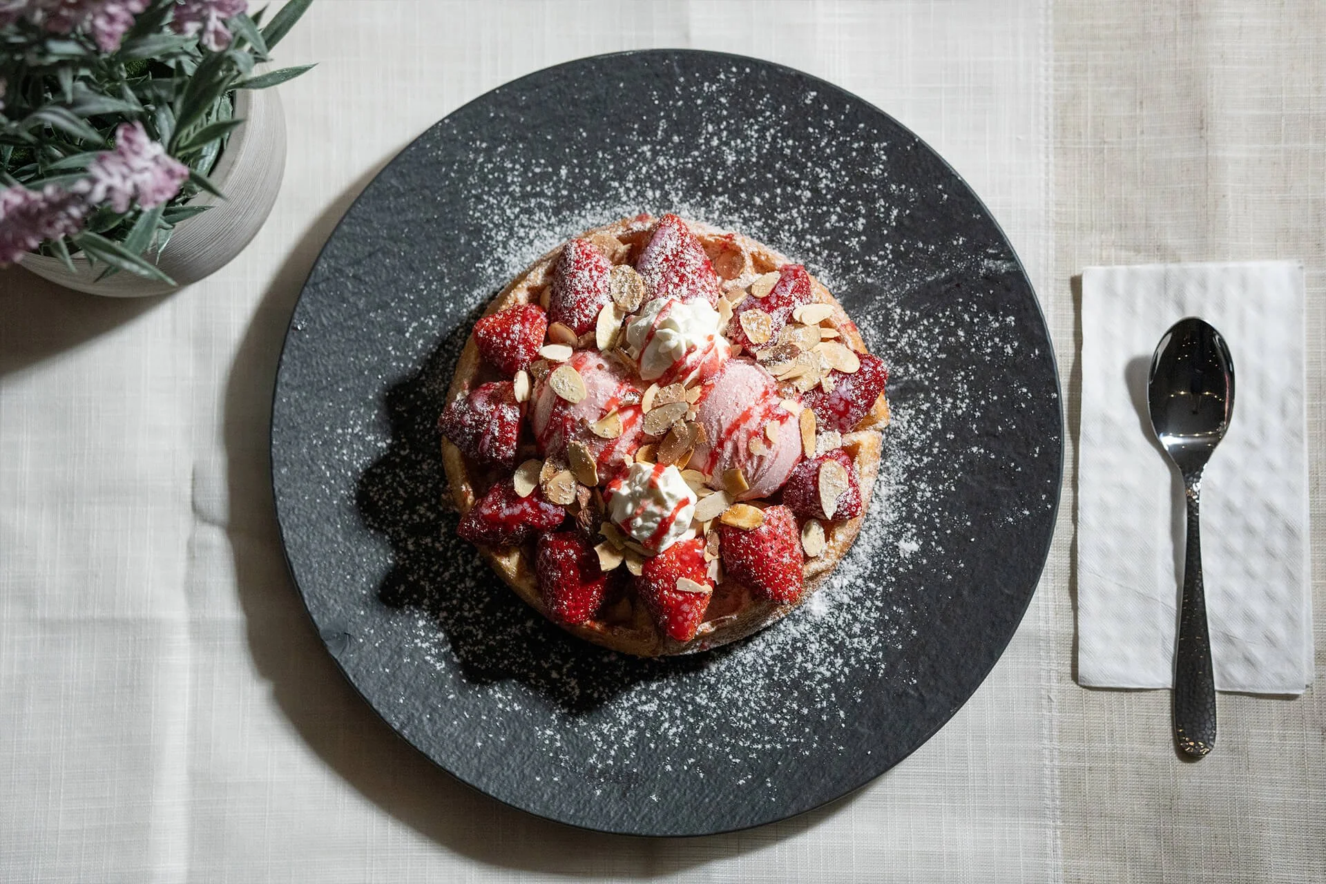 A waffle topped with strawberries, whipped cream, sliced almonds, and powdered sugar on a black plate. A spoon is placed on a white napkin to the right, and a vase with purple and green flowers is on the left, all on a light-colored table.