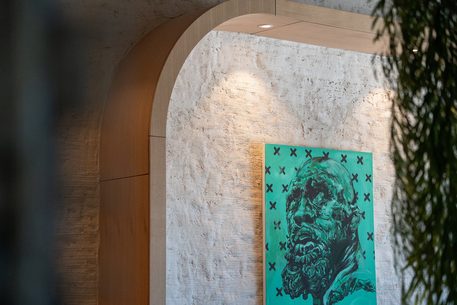 A gallery wall featuring an abstract portrait painting of a bearded man with long hair, set against a green background with black X marks, illuminated by wall-mounted lights on a textured white wall, with a wooden arch and some greenery in the foregr