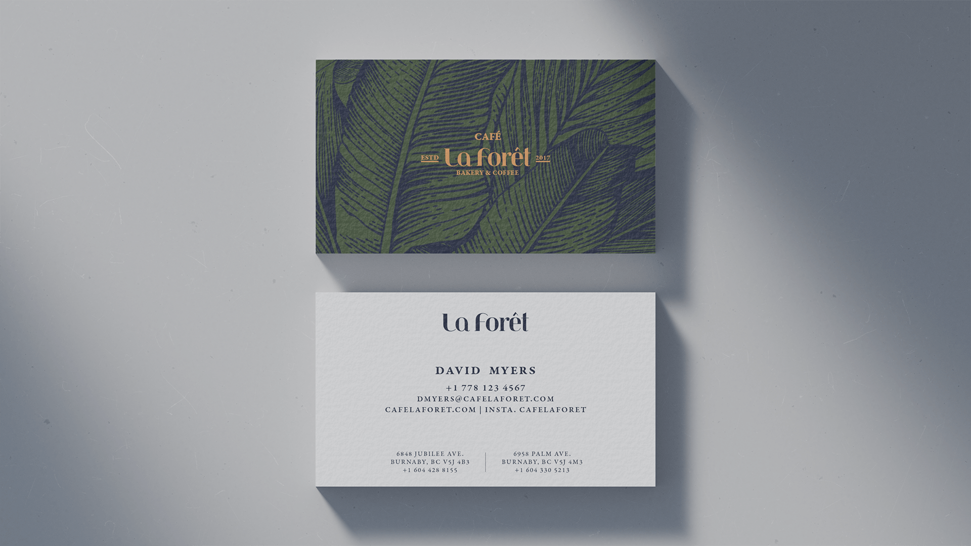 A business card with the text 'La Forêt', providing contact details including an email, phone number, and addresses for two locations. A matching invitation or advertisement with a green botanical print and gold text is placed above it.
