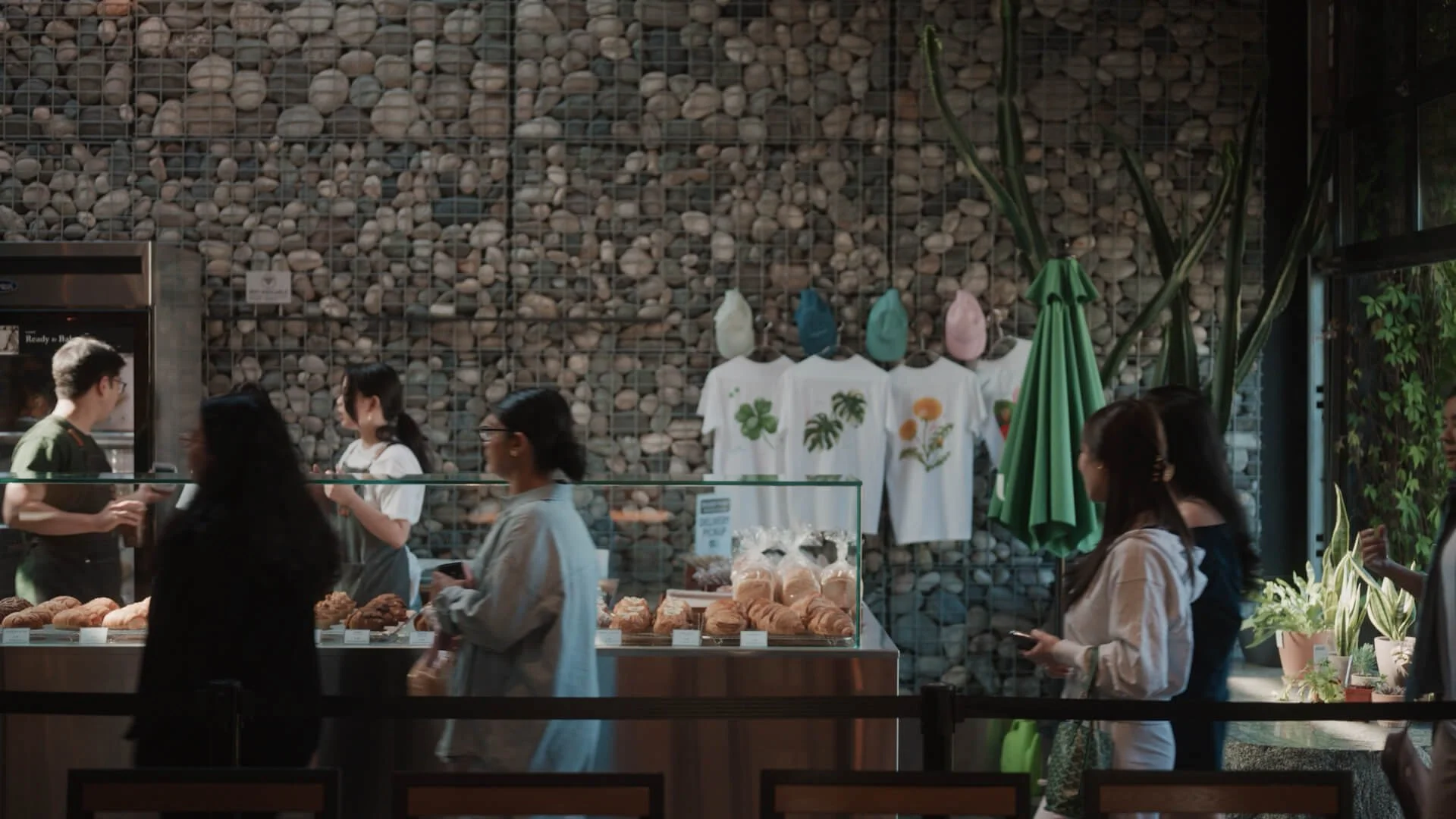 People shopping and chatting inside a bakery or cafe with a stone wall background, t-shirts on display, and baked goods in the foreground.
