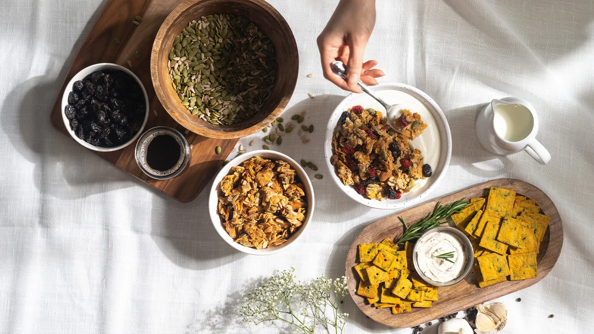 A table with vegan food including a bowl of granola with berries, a bowl of granola with pumpkin seeds, a bowl of dried fruit, a small pitcher of milk, a platter of sliced pumpkin savory squares with herbs, and a small bowl of yogurt garnished with h