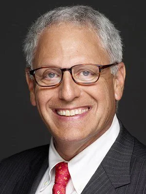 Close-up of a smiling man with gray hair and glasses, wearing a gray suit, white shirt, and red patterned tie, against a dark background.