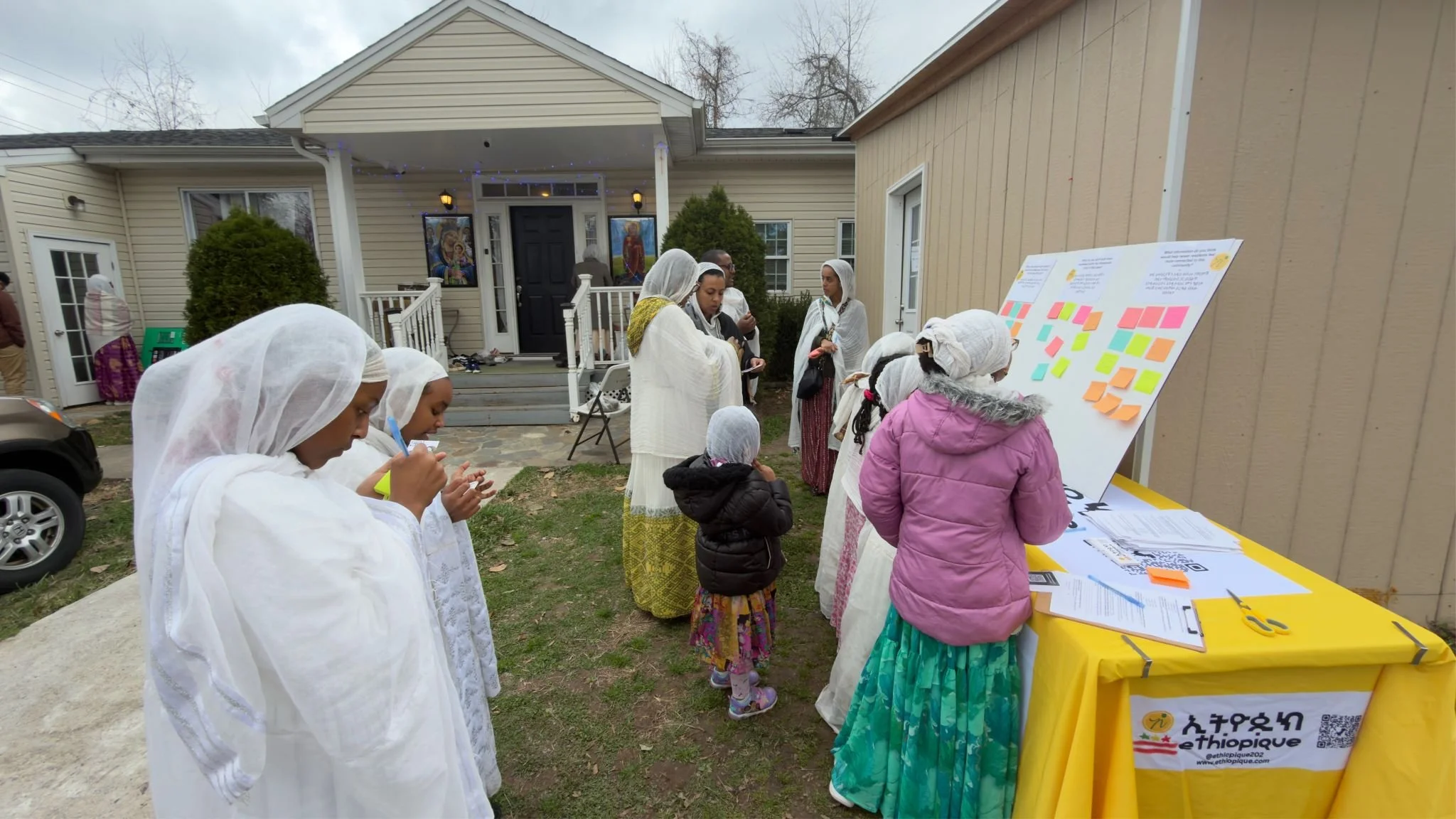 People gathered outdoors in front of a house, engaged in cultural or community event activities. Some are wearing traditional Ethiopian clothing, and a table displays informational materials, while a board with sticky notes is visible. The scene appears to be a multicultural or religious gathering.
