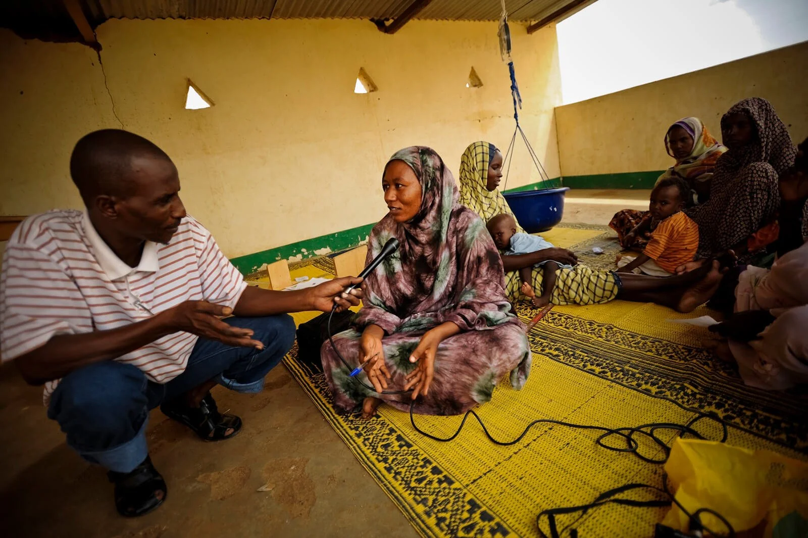 A group of women and children sitting on a yellow woven mat inside a simple room with yellow walls. A woman in the center is being interviewed by a man holding a microphone.