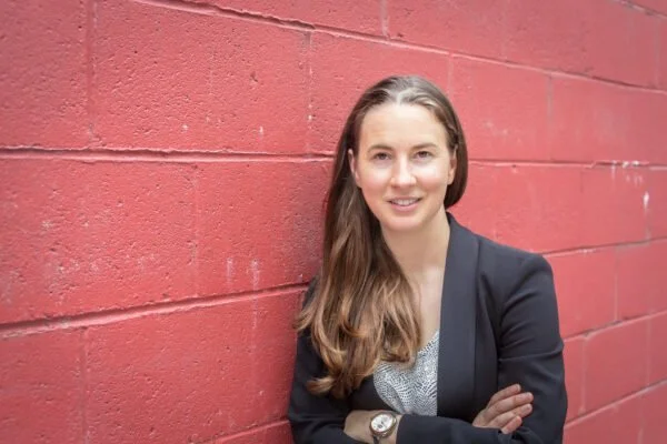 A woman with long brown hair, wearing a black blazer and gray shirt, standing with arms crossed against a red brick wall.