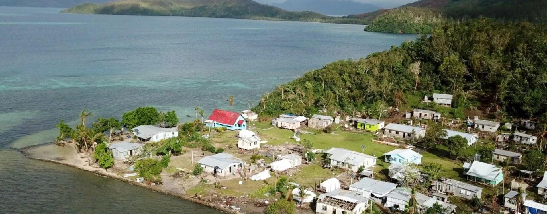 Aerial view of a small coastal village with colorful houses, surrounded by lush green trees, next to a body of water with a forested hillside in the background.
