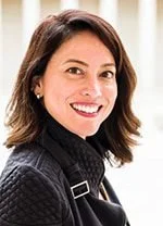 A woman with shoulder-length dark hair smiling, wearing a dark sweater and a black strap over her shoulder.
