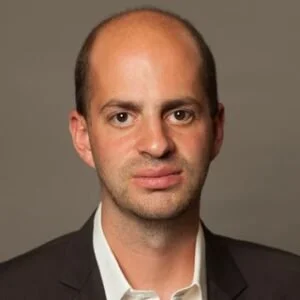 A portrait of a man with short hair, wearing a dark blazer and a white shirt against a neutral background.