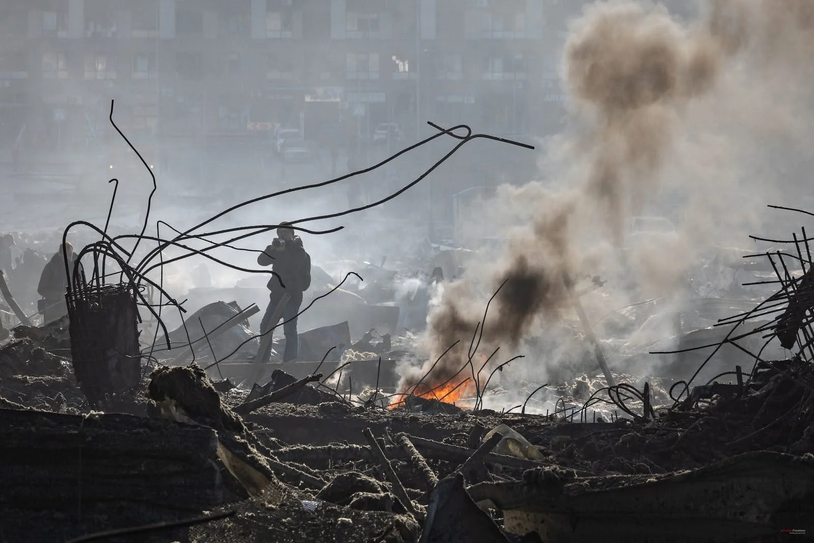 Debris and destruction with smoke rising from a fire, and people in the background amid chaos.