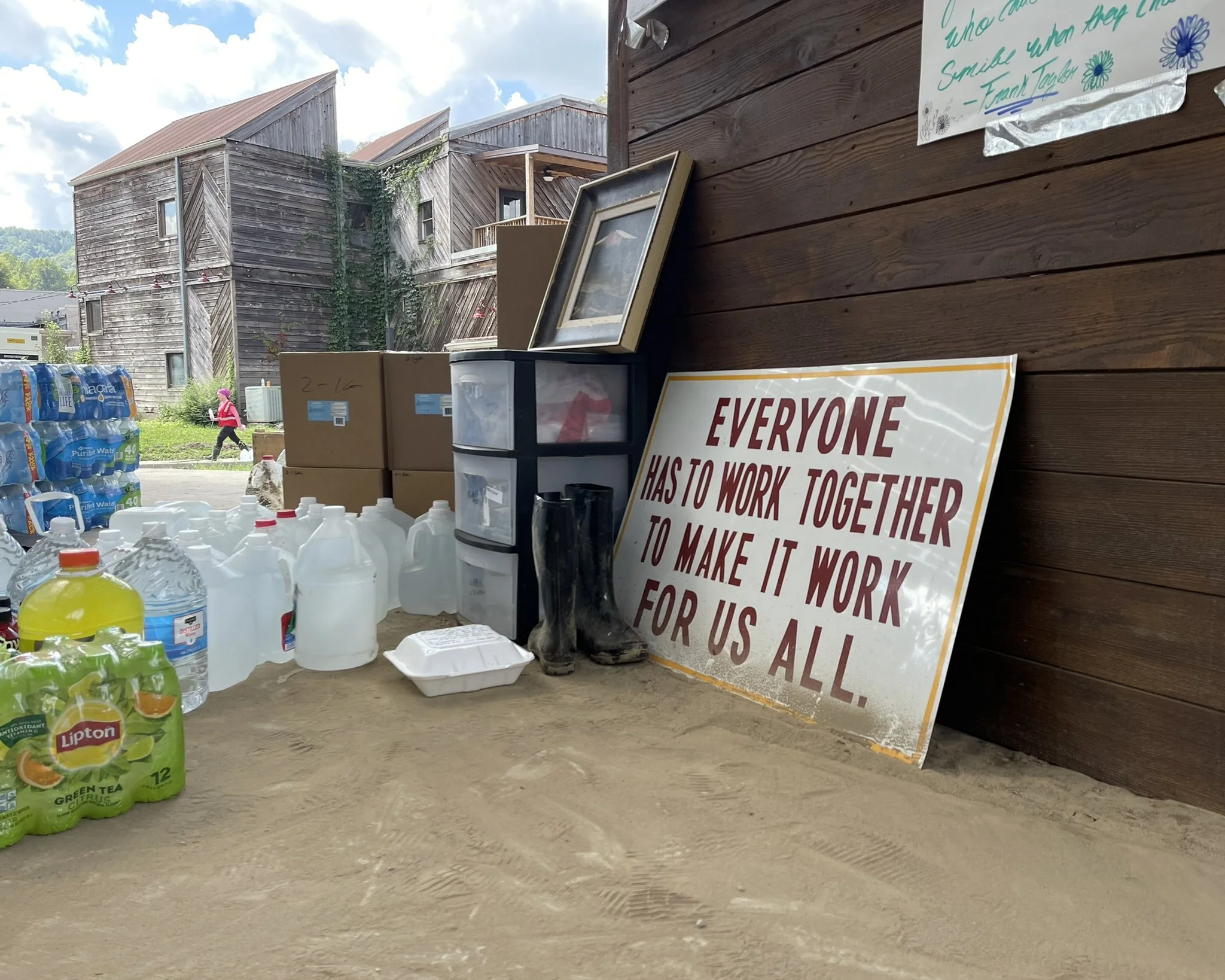 View of stacked bottled water, paper towels, and cardboard boxes on a concrete ground, with a wooden building and cloudy sky in the background. A sign in the foreground reads, 'Everyone has to work together to make it work for us all.'