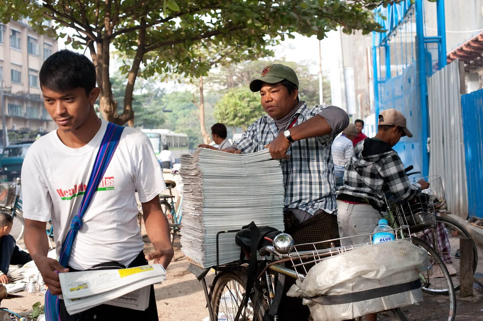 A man on a bicycle selling newspapers on a busy street.