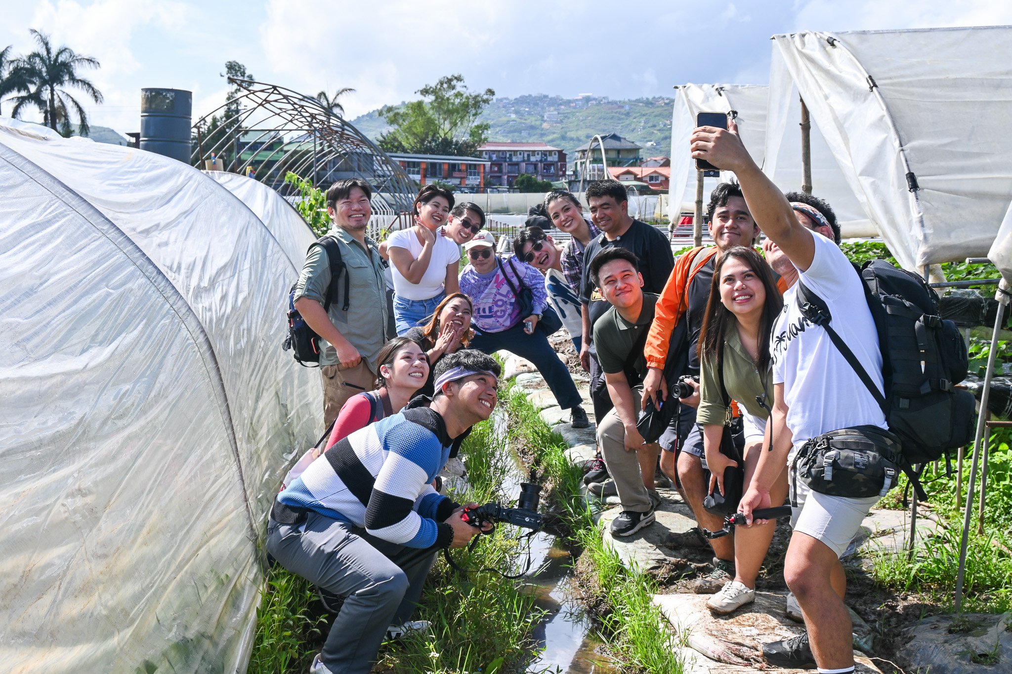 Group of people taking a selfie outdoors in a garden or farm, with greenhouses and hills in the background.