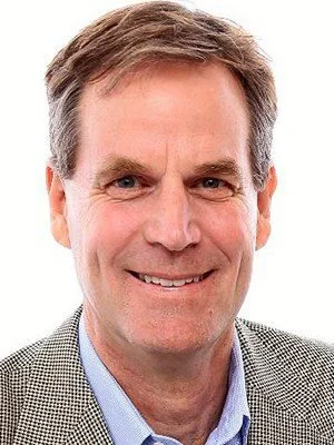 Close-up of a smiling middle-aged man with brown hair, wearing a gray blazer and blue collared shirt, against a white background.