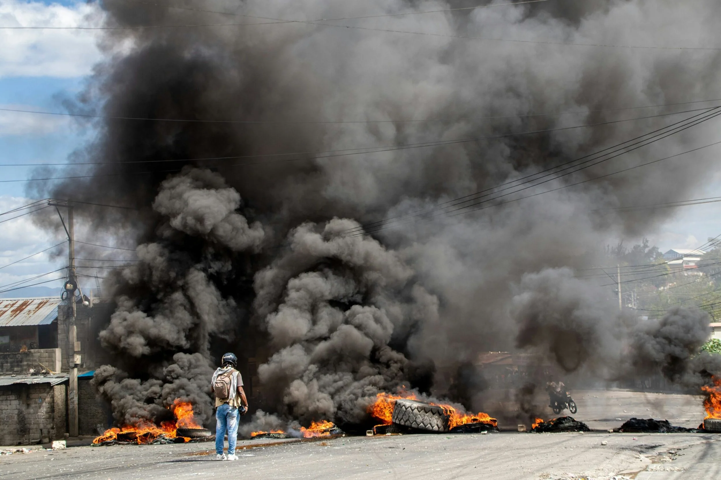 A person walking away from a large fire with thick black smoke and flames on the street, with burning tires and debris.
