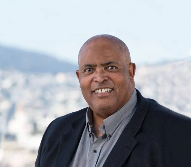 A man with a shaved head, dark dress shirt, and blazer smiling outdoors with a cityscape and hills in the background.