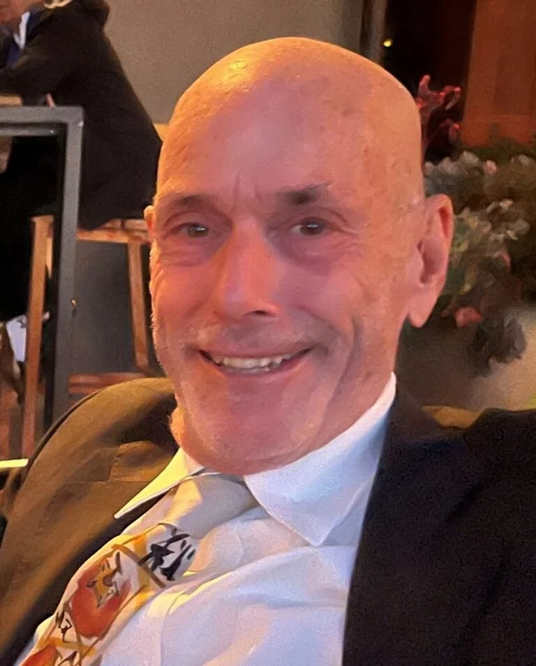 A smiling, bald man in a suit and tie sitting indoors with flowers and other people in the background.