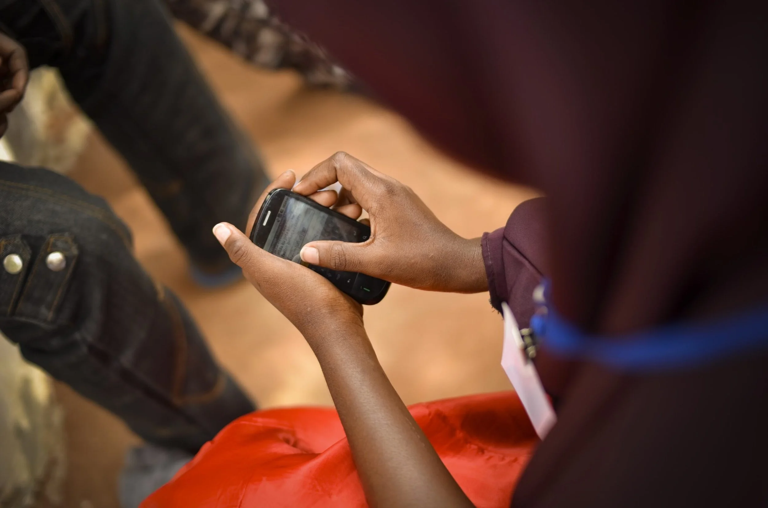 A person with dark skin is holding a smartphone, browsing or typing on it, while sitting down. They are wearing a maroon long sleeve shirt and bright red pants. The background shows a person sitting with their legs crossed, wearing black boots and jeans.