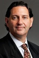 A professional man with dark hair, wearing a dark suit, white shirt, and patterned tie, looking directly at the camera.