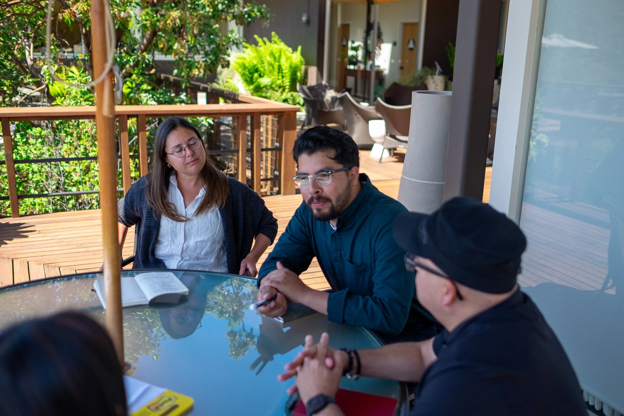 Group of five people sitting around a glass table outdoors, engaged in a discussion, with open notebooks and pens on the table.