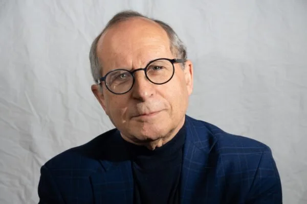 A man with glasses wearing a dark checkered blazer and black turtleneck, posing against a white backdrop.